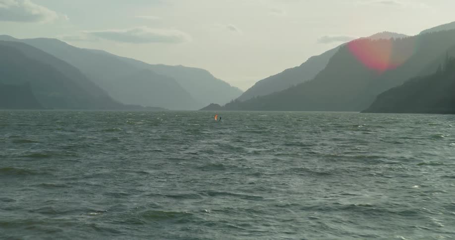 A solitary wing foiler glides across the choppy river waters of a vast gorge, dwarfed by the majestic, hazy mountains in the background
