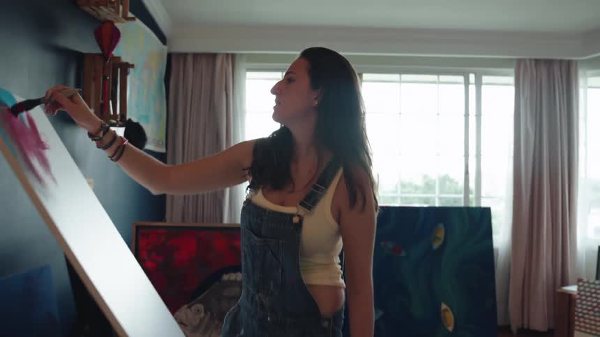 A young female artist passionately applies broad strokes of pink paint to a large canvas in her sunlit studio