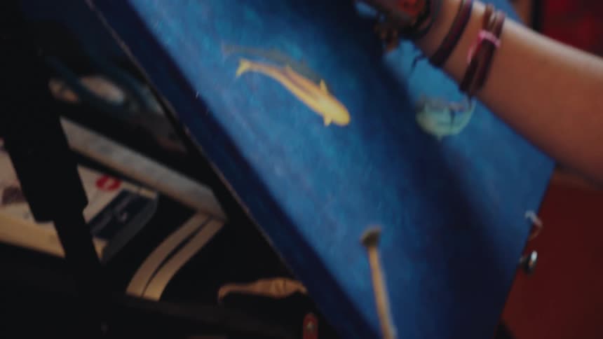 A slow tilt up from messy art supplies reveals an artist's hand carefully painting fish onto a canvas