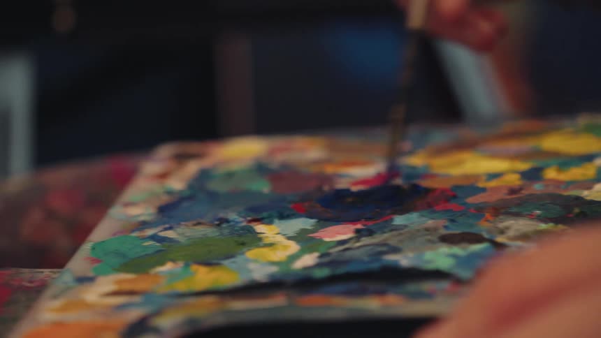 A close-up reveals an artist mixing blue paint on a vibrant palette