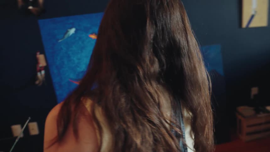 Camera pulls back from a focused female artist painting colorful fish on a large blue canvas in her creative home studio