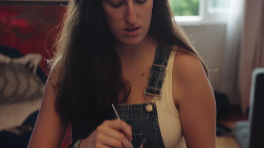 A focused female artist in overalls selects a paintbrush from a colorful pot and concentrates on her work in a naturally lit studio