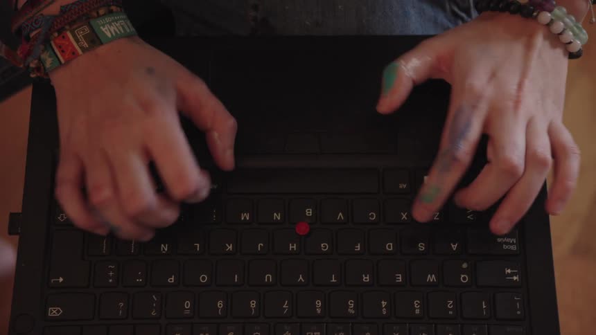An artist's hands, stained with paint, type on a laptop keyboard in a top-down close-up view