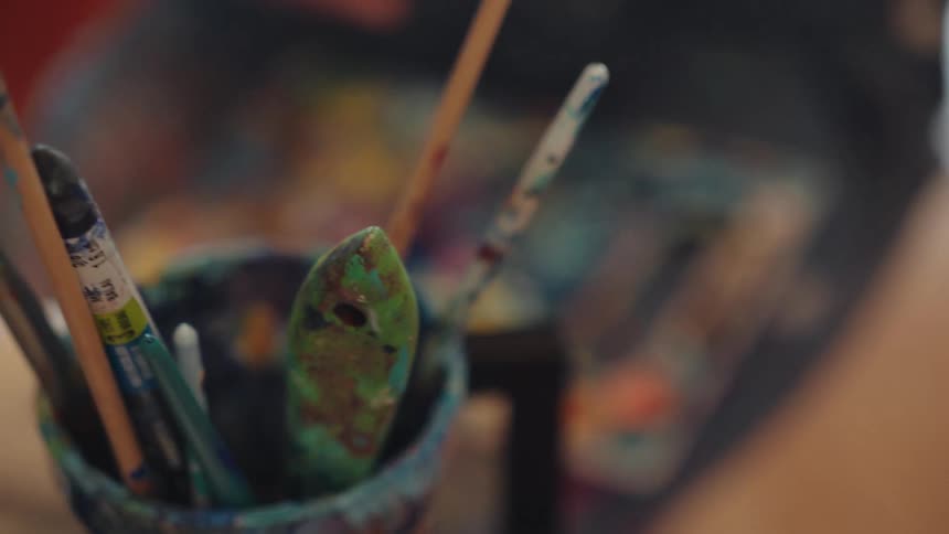 A close-up view of an artist mixing paint on a colorful palette, with the focus shifting to a jar of used paintbrushes