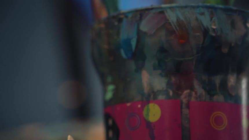 Close up of an artist's hand painting a canvas, rack focusing to a cup full of used paintbrushes