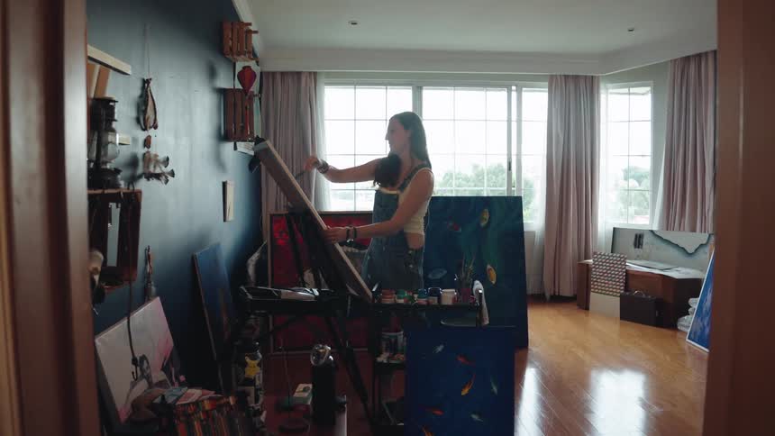 A young woman paints on a canvas at her easel in a sunlit art studio filled with her creations
