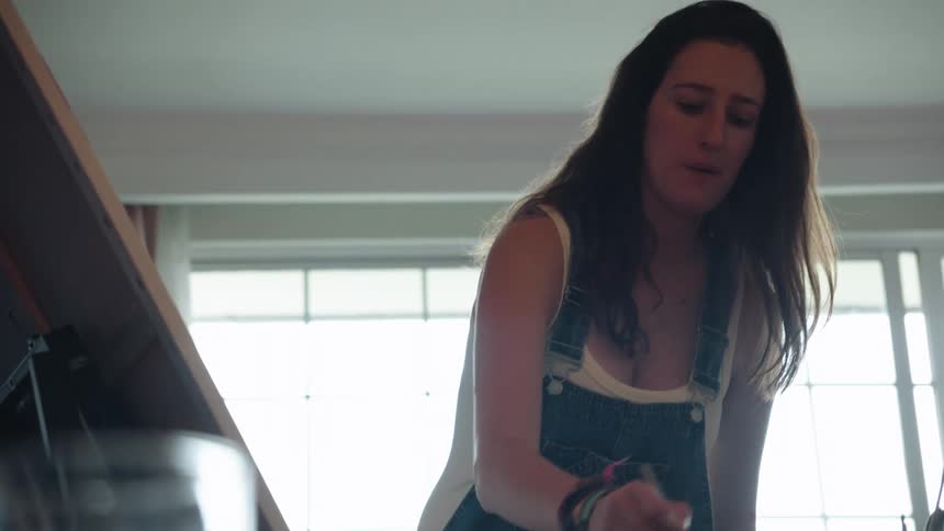 Rack focus from a glass of water reveals a female artist in overalls painting on a canvas in her studio in slow motion