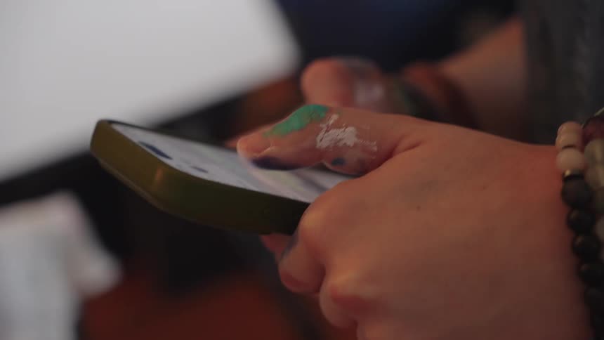 An artist in denim overalls with paint-stained hands takes a break to scroll on their smartphone