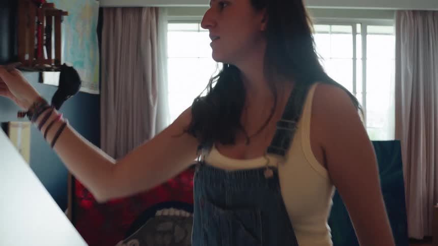 Female artist in overalls paints on a large canvas in her bright home studio, with a push-in to a close-up of the brushstrokes