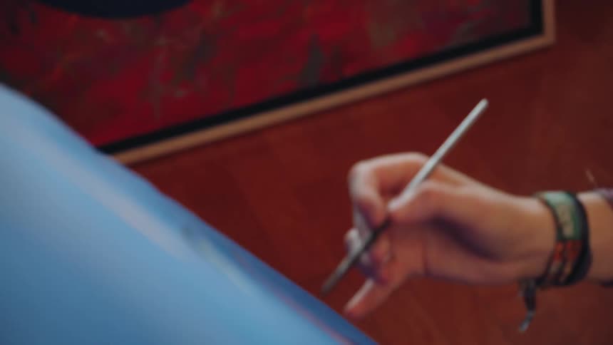 A close-up of a thoughtful young female artist tilts down to reveal her hand painting on a large blue canvas in her sunlit studio