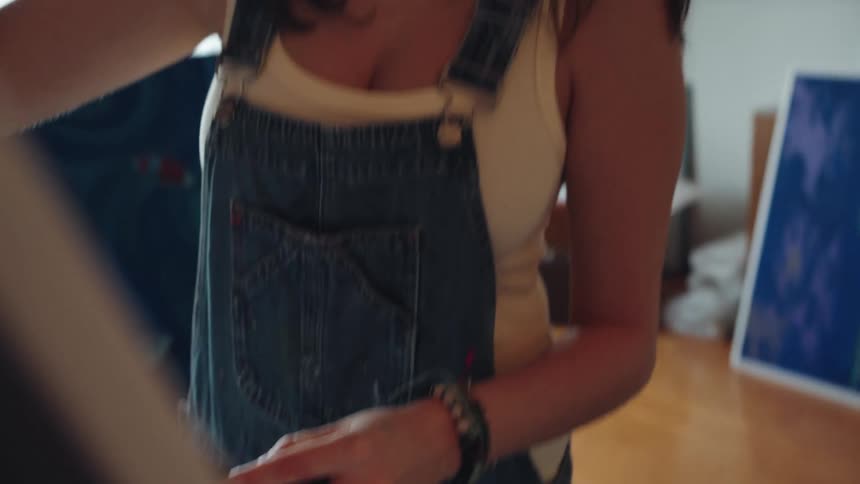 Close-up slow-motion reveal of a focused young female artist in overalls painting on a large canvas in her sunlit studio