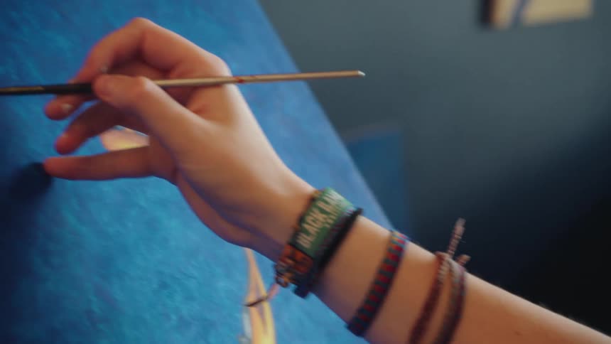 A female artist, wearing colorful bracelets, carefully paints the fine details of a fish on a blue canvas, revealing her focused profile in slow motion