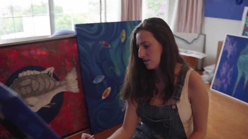 A young female painter works on a new piece in her art studio, surrounded by her colorful finished canvases