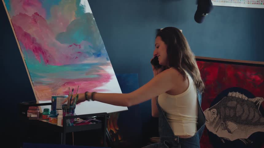 A young female artist smiles while talking on her phone in a creative studio next to her colorful abstract painting