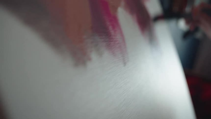 A female artist in denim overalls paints abstract strokes of pink onto a blank canvas in her sunlit home studio