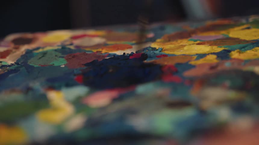 An artist's brush mixes dark blue paint on a messy, colorful palette in an intimate close-up shot