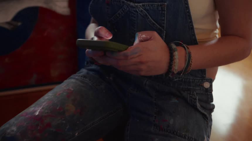 A young female artist takes a break from her painting to scroll through her smartphone in a sunlit art studio