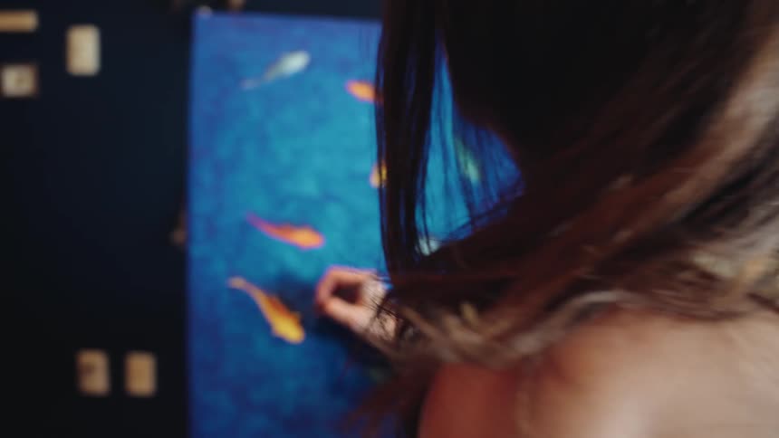 A young female artist adds the finishing touches to her vibrant painting of koi fish swimming in her cozy home studio