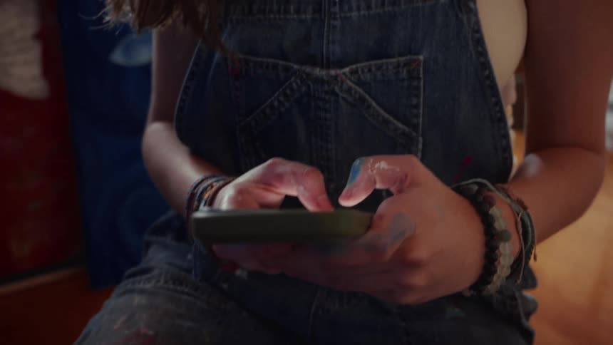 A young female artist in paint-stained overalls takes a break in her studio, using her phone before looking up to stare directly into the camera
