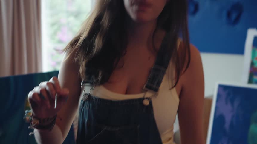 Slow motion reveals a focused female artist in overalls, holding a paintbrush and contemplating her work in a bright studio