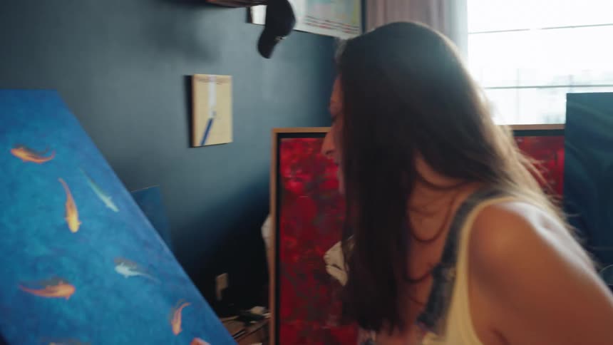 A focused female artist sits in her home studio, carefully adding details to a vibrant painting of colorful fish on a blue canvas