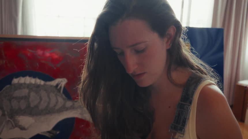 A female artist wearing overalls in her sunlit studio takes a break from painting to answer a phone call, her expression serious