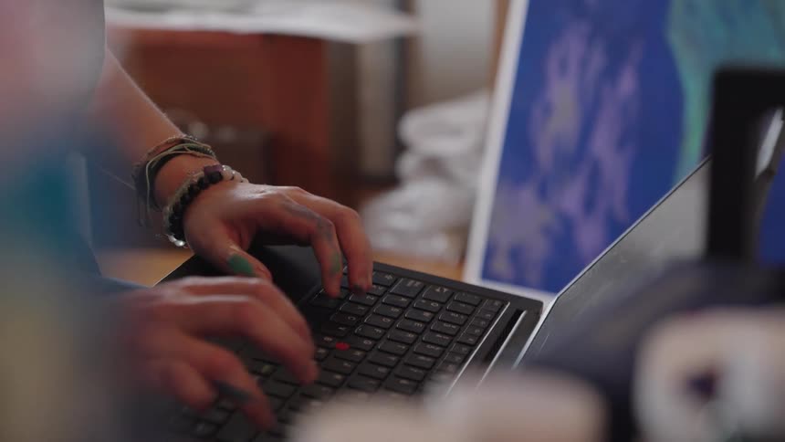 A young female artist with paint-covered hands wearing overalls works on her laptop in a creative art studio