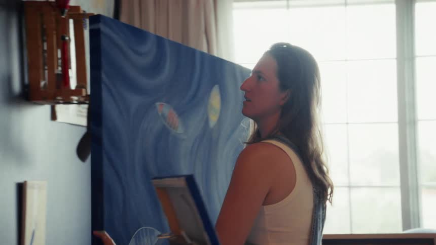 A barefoot female artist in overalls picks up her large abstract blue painting from the floor and hangs it on the wall of her studio in slow motion
