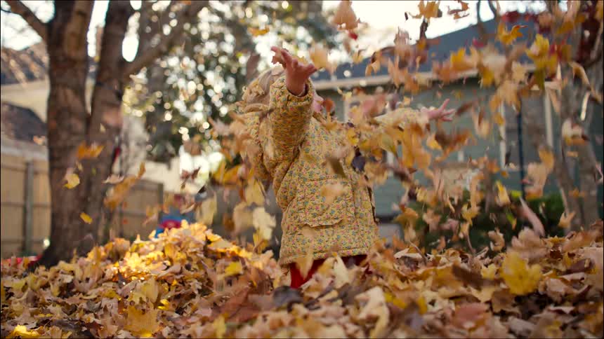 A little girl stands up from a pile of leaves and throws them into the air in slow motion in her backyard