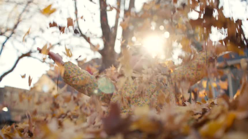 A joyful young girl rises up from a pile of colorful autumn leaves, tossing them into the air in slow motion as the sun shines