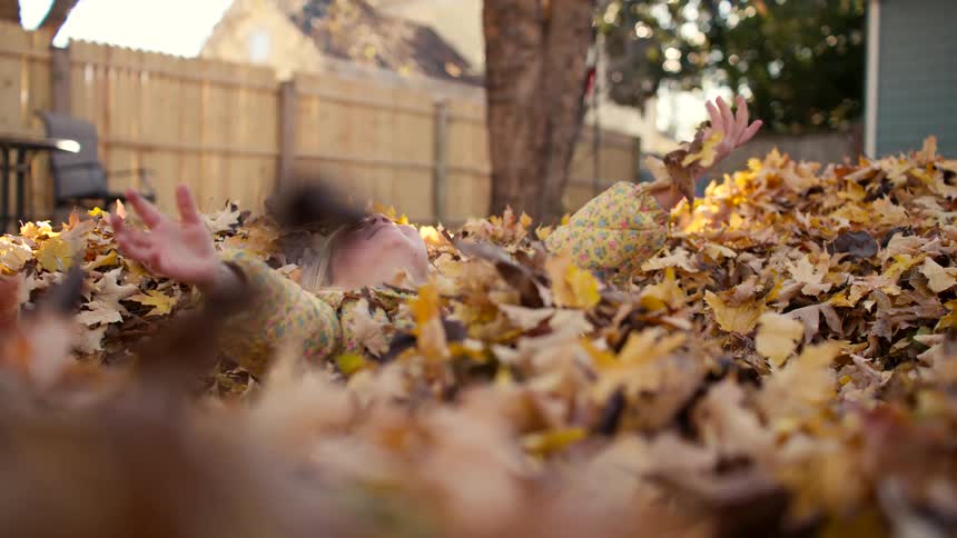A joyful little girl peeks out from a giant pile of autumn leaves before falling back and throwing them into the air in super slow motion