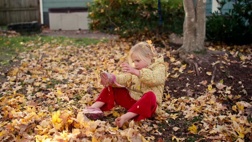 A cute blonde girl in a yellow coat sits barefoot in a pile of fall leaves and tries to shake them out of her shoe in slow motion