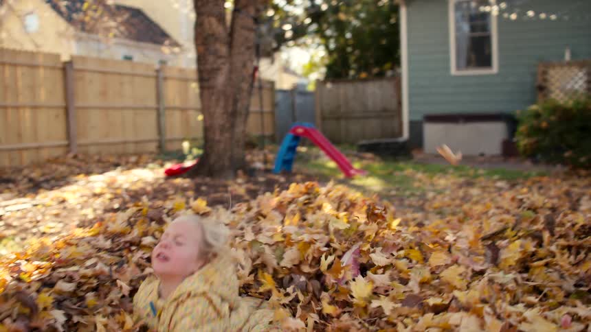 A joyful little girl in a yellow coat runs in slow motion and jumps headfirst into a large pile of autumn leaves in her backyard