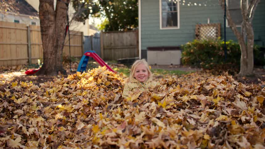 A happy little girl runs in slow motion and joyfully jumps into a big pile of fallen autumn leaves in her backyard