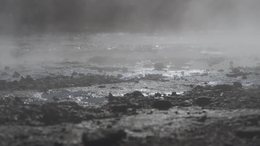 Backlit steam rises from the dark, rocky ground of a volcanic geothermal field
