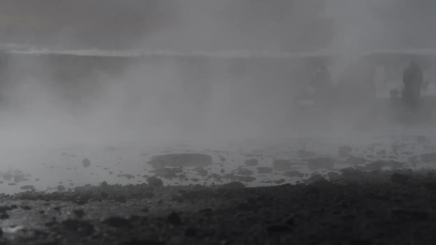 Thick, mysterious steam rises from a rocky geothermal hot spring, with faint silhouettes of people visible in the distance