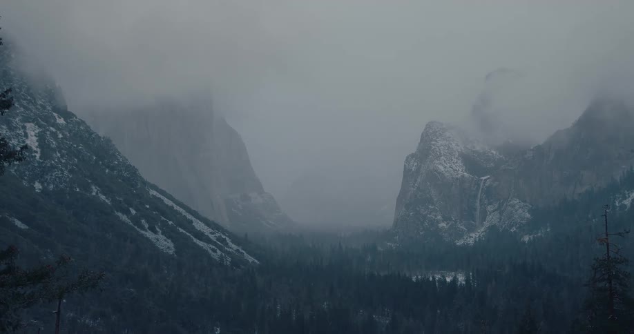A moody, wide shot of the iconic Yosemite Valley as snow falls on the mountains and forest during a foggy winter day
