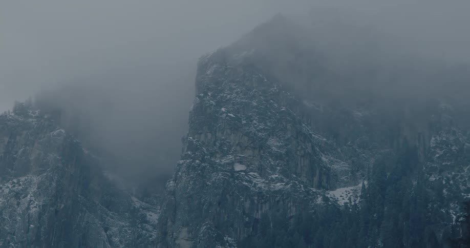Thick fog drifts slowly across a snow-covered mountain, creating a moody and atmospheric winter scene