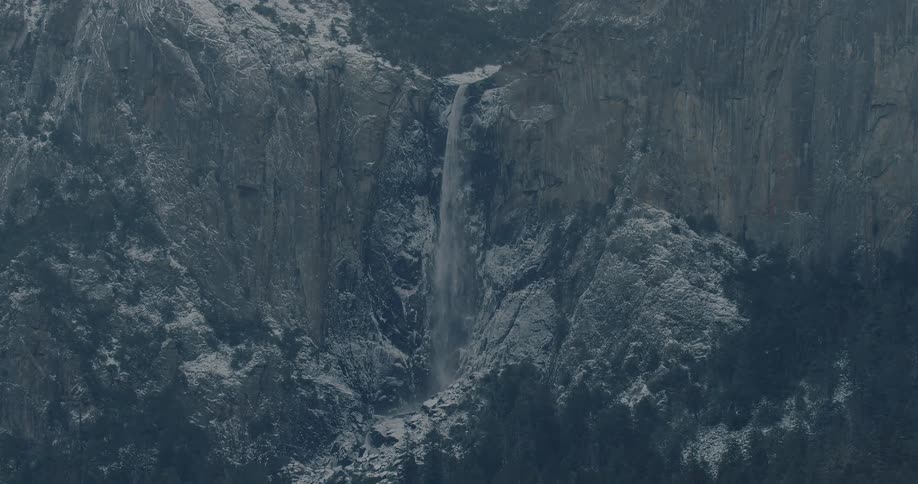 A powerful waterfall cascades down a sheer, snow-dusted rock face in a vast mountain landscape