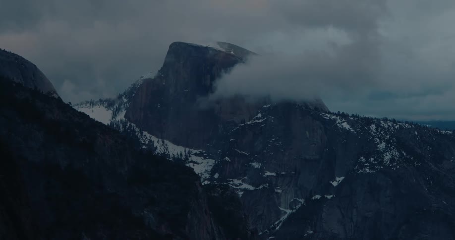 A dark and moody shot of the iconic Half Dome mountain in Yosemite National Park shrouded in slow-moving clouds at dusk