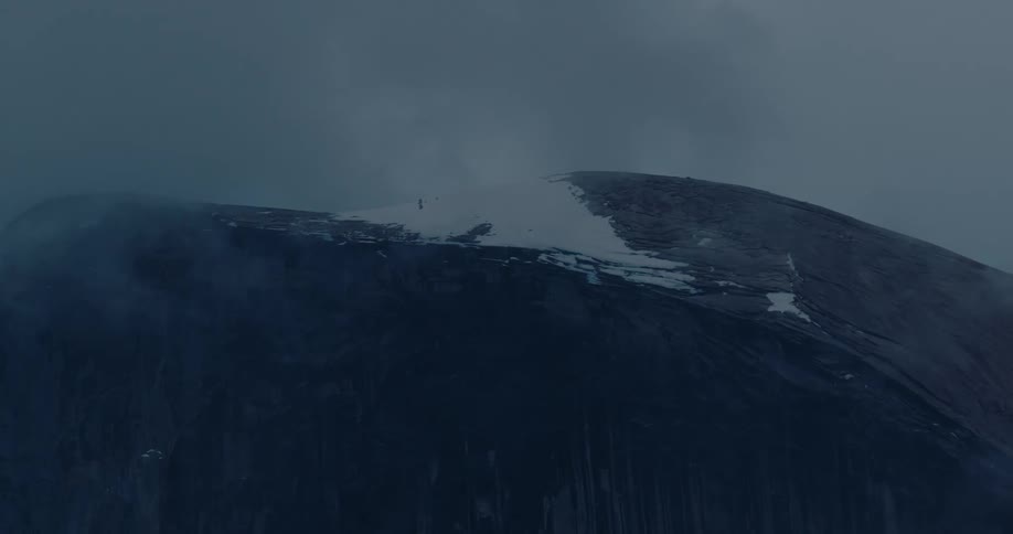 Tiny climbers ascend a massive, snow-capped volcano through thick fog under a dark and moody sky
