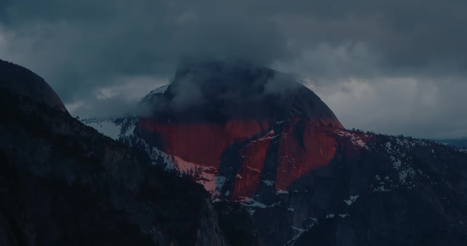 Dramatic alpenglow illuminates the face of a mountain peak shrouded in dark, moody clouds at sunset