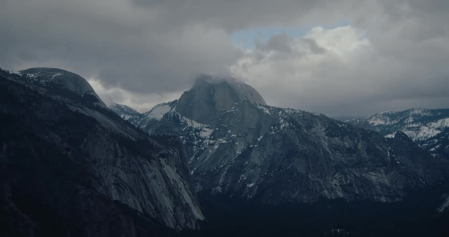 A moody and dramatic wide shot of a snow-capped mountain range partially hidden by dark, overcast clouds