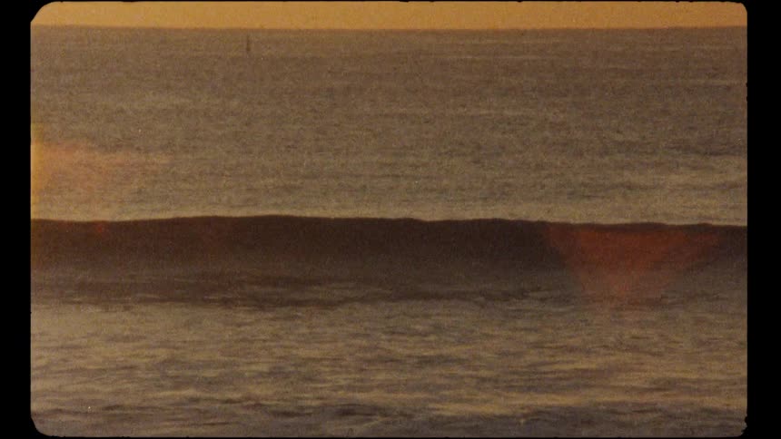 A solitary ocean wave rolls across the water during a warm, hazy sunset in this vintage film-style footage