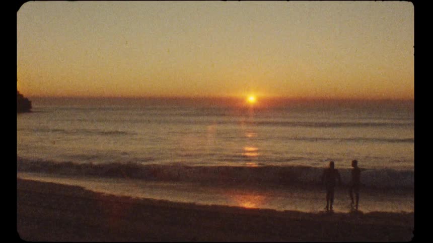A nostalgic, grainy shot shows two people in silhouette enjoying a serene sunset on a tranquil beach