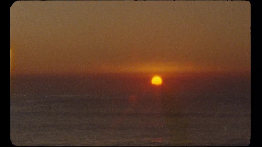 A grainy, vintage Super 8 film captures a beautiful orange sunset over the calm ocean horizon