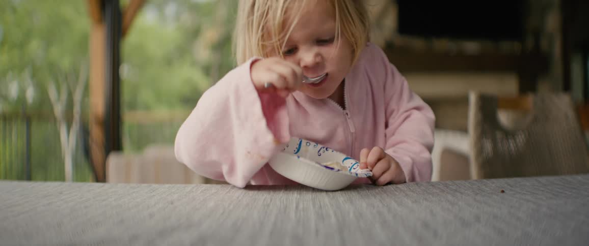 A close-up slow-motion shot shows a young blonde girl in a pink jacket eating from a bowl with a spoon