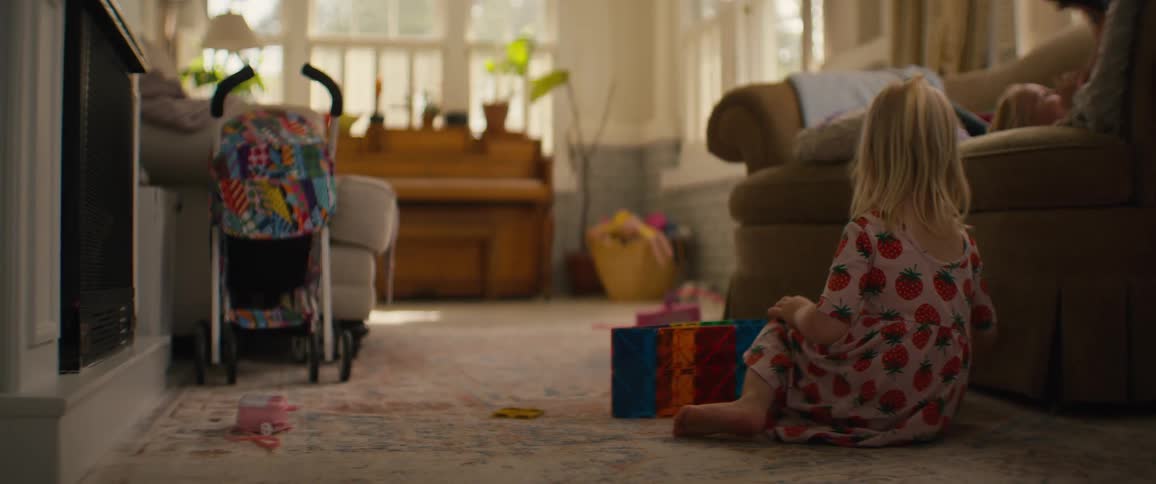 A young girl in a strawberry-print dress plays with colorful magnetic building tiles on the living room floor