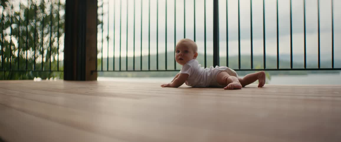 Cute baby in a white onesie lies on a wooden porch deck, looking back at the camera with a lake in the background