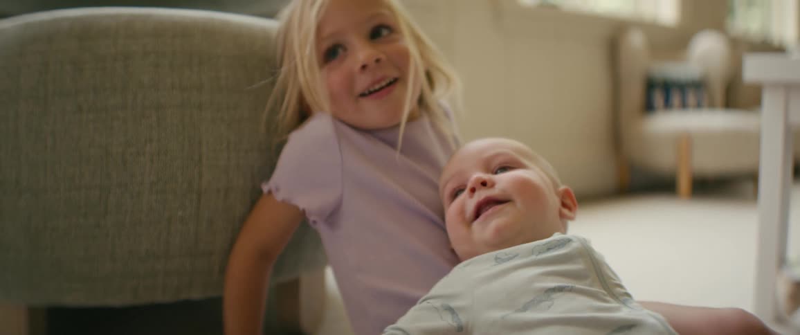 A happy baby boy laughs with pure joy while his older sister holds him in a sweet sibling moment